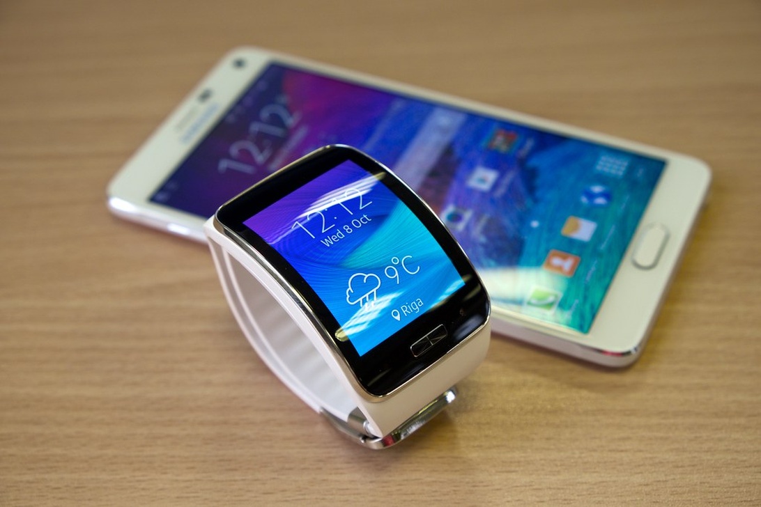 En silversmartklocka med vitt armband visar väderinformation (9°C) framför en vit Samsung-smartphone, båda med liknande blå gränssnitt på en träyta, vilket tydligt visar deras anslutna relation.