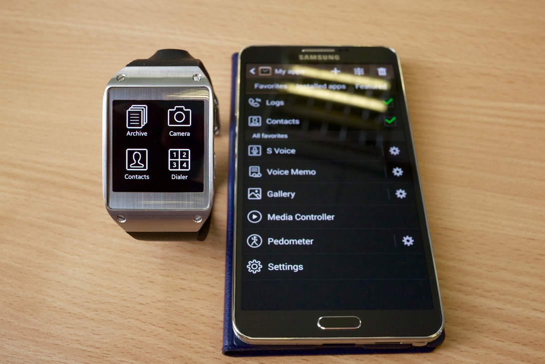 En Samsung Galaxy Gear smartwatch med appikoner (Arkiv, Kamera, Kontakter, Dialer) bredvid en Samsung-smartphone som visar tillhörande appgränssnitt med anslutna funktioner och inställningsmeny på en träyta.
